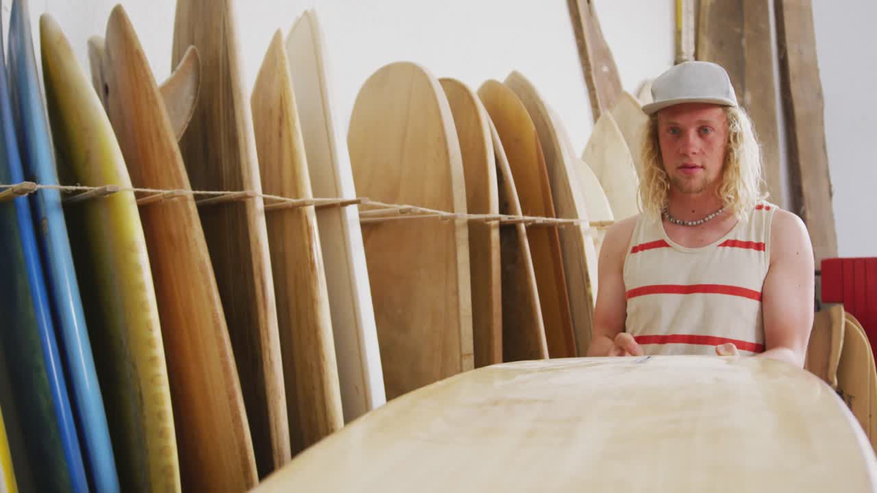 백인 남성 서프보드 제작자가 스튜디오에서 서프보드 중 하나를 확인하고 있습니다.