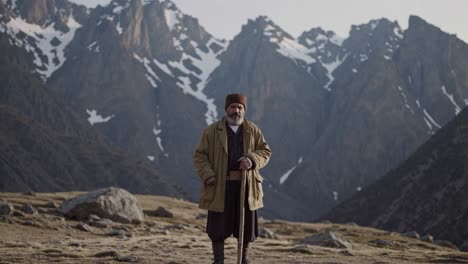 elderly shepherd wearing traditional clothing stands proudly in a desolate mountain valley, holding a wooden staff with majestic snow capped peaks in the background