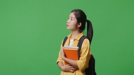 side view of asian woman student with a backpack and some books smiling and looking around while standing in the green screen background studio