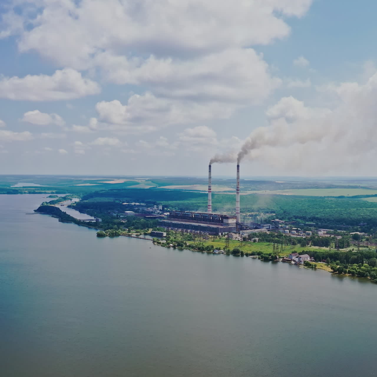 Beautiful scenery of the river background and plant with pipes. Natural landscape of blue water, green fields and factory nearby. Aerial view. Motion up.