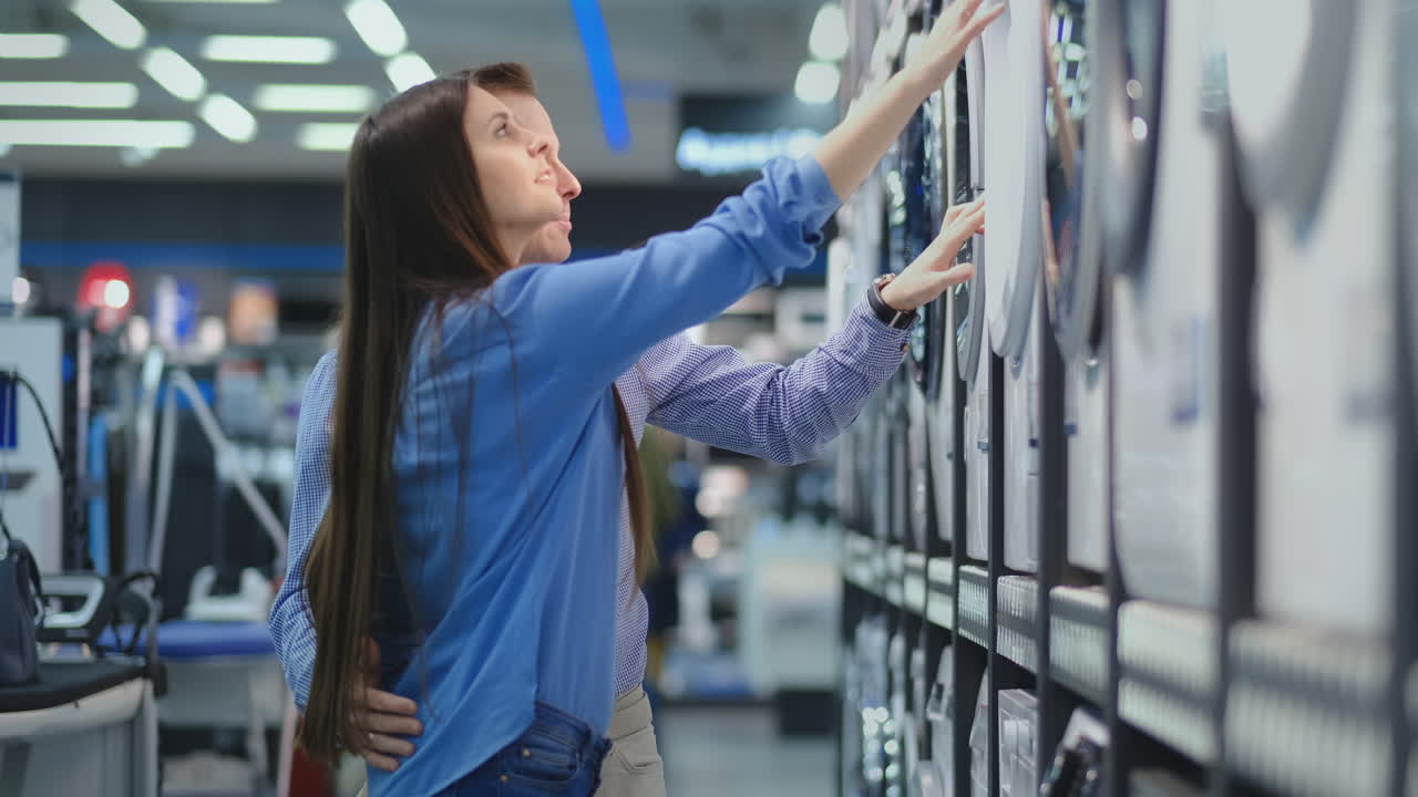 Young man and woman in appliances store choose to buy washing machine for home. Open the door looking into the drum, compare the design and characteristics of the devices.