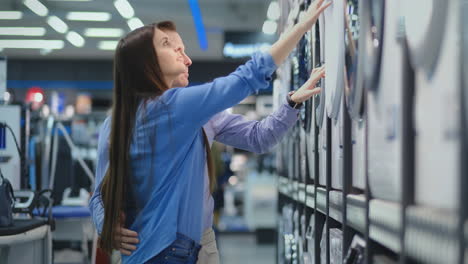 young man and woman in appliances store choose to buy washing machine for home. open the door looking into the drum, compare the design and characteristics of the devices.