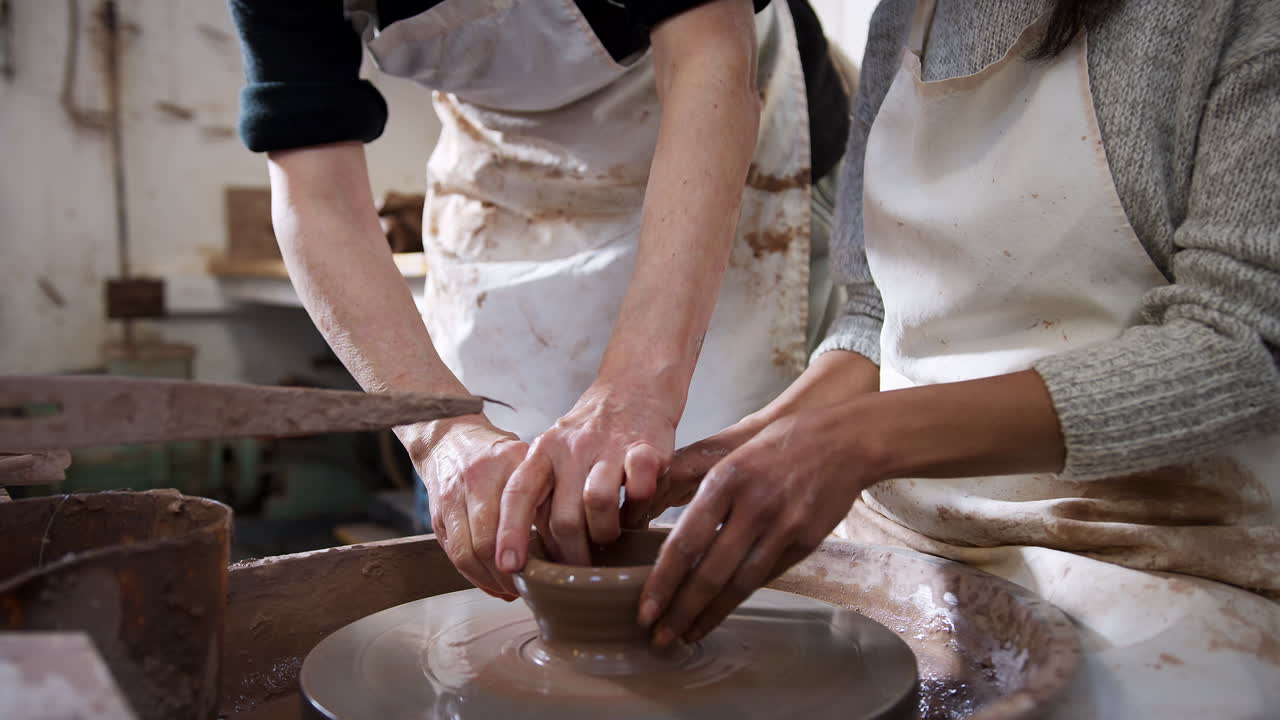Close Up Of Male Teacher Helping Woman Sitting At Wheel In Pottery Class