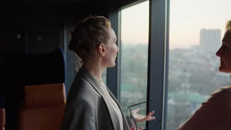 una confiada mujer de negocios de mediana edad en un traje de negocios gris se comunica con su colega mientras está de pie cerca de ventanas panorámicas con vistas a la ciudad en una oficina moderna