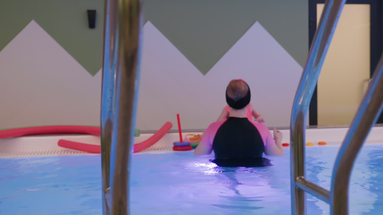 검은색과 핑크색 바지를 입은 여성이 수영장 옆에서 핑크색 수영복 바지를 입은 어린 소녀와 놀고 있는 뒷모습, 부드러운 상호 작용, 따뜻한 빛, 가족 간의 유대감, 즐거운 코칭, 안전한 환경, 솔직한 순간