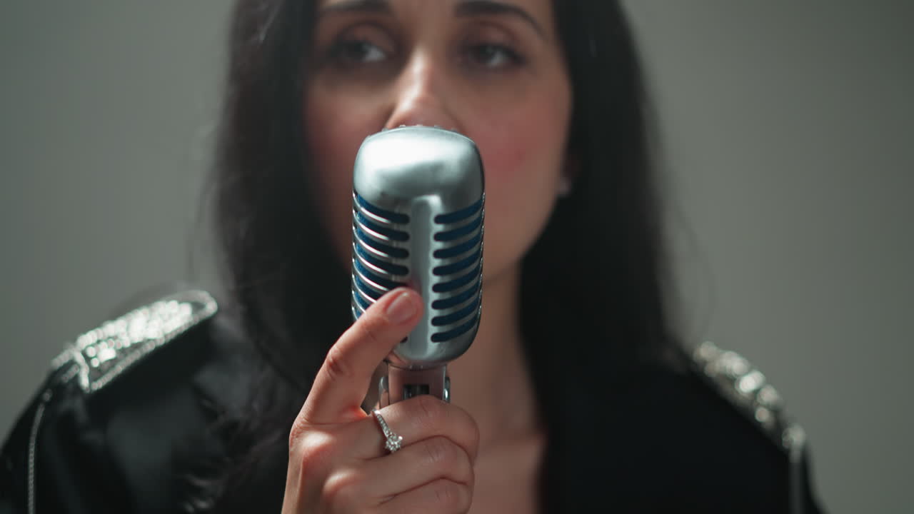 primer plano de una cantante femenina seria en un traje negro, agarrando un micrófono vintage con una mano, mientras la cámara hace zoom contra un fondo borroso