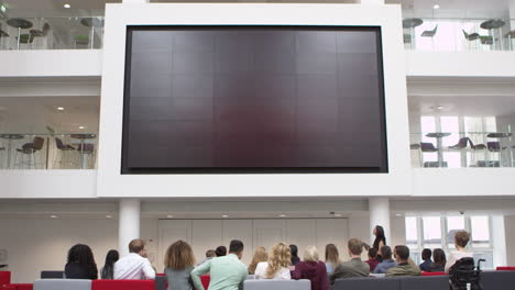 estudiantes en una conferencia viendo una pantalla grande en un atrio de la universidad, filmado en r3d