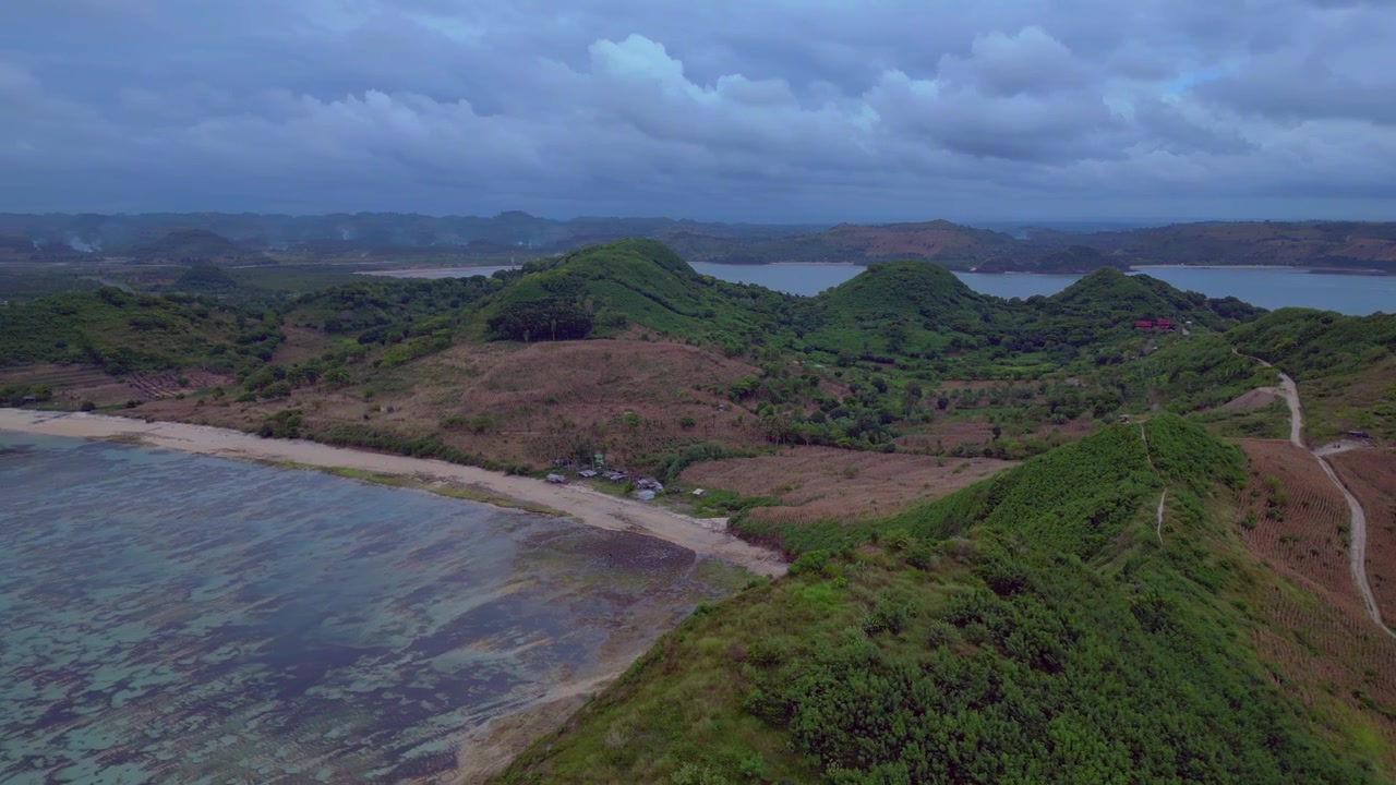 Scoprire l'isola tropicale di Lombok con vaste colline coperte da una fitta vegetazione verde che incontrano le calme acque della baia sotto un cielo nuvoloso. Incredibile vista aerea con rotazione panoramica a sinistra da drone.