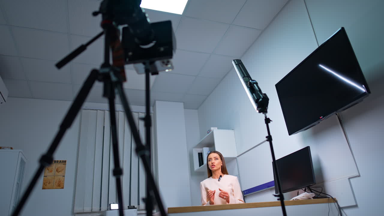 Camera on tripod filming a woman sitting at desk. Female Caucasian blogger talks and gestures with her hands. Low angle view.