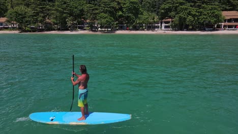 Vista-De-Pájaro-De-Drones-Aéreos-De-Camiones-De-ángulo-Bajo-De-Un-Hombre-Caucásico-Haciendo-Ejercicio-En-Una-Tabla-De-Remo-En-Aguas-Turquesas-Tropicales-Claras,-Con-Resort-De-Playa-Y-Costa-En-Tailandia