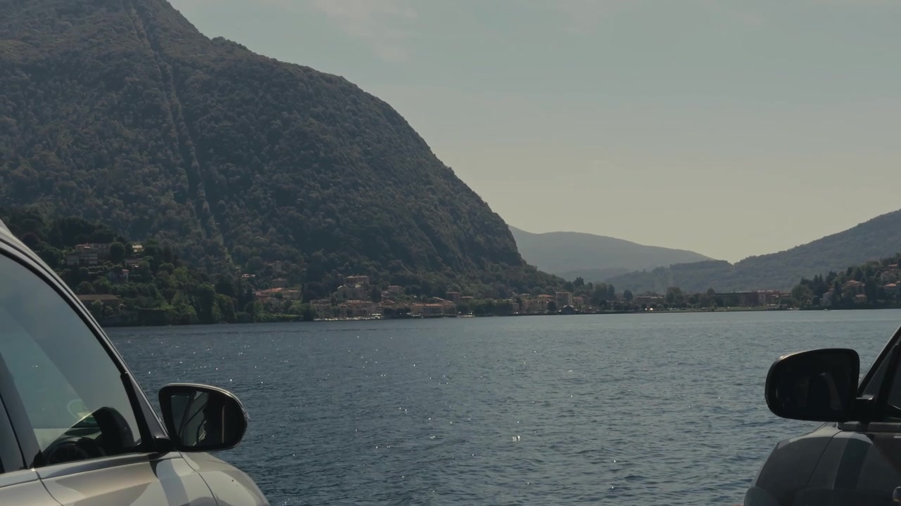 Punto di vista creativo da un traghetto che guarda tra due specchietti retrovisori di auto verso l'acqua calma del lago e spettacolari montagne alpine, catturando un suggestivo attraversamento in traghetto italiano in una giornata estiva.