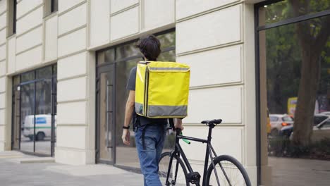 Delivery-man-walking-with-bike-and-yellow-bag,-looking-for-address,-rear-view