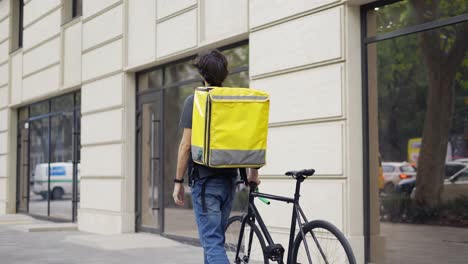 hombre de entrega caminando con bicicleta y bolsa amarilla, buscando dirección, vista trasera