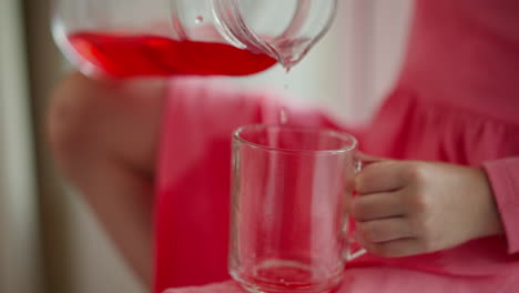 hand view of child pouring red juice from glass jar into clear cup, part of drink spilled on pink dress, small fingers gripping mug handle, bright daylight from window and blurred background of room