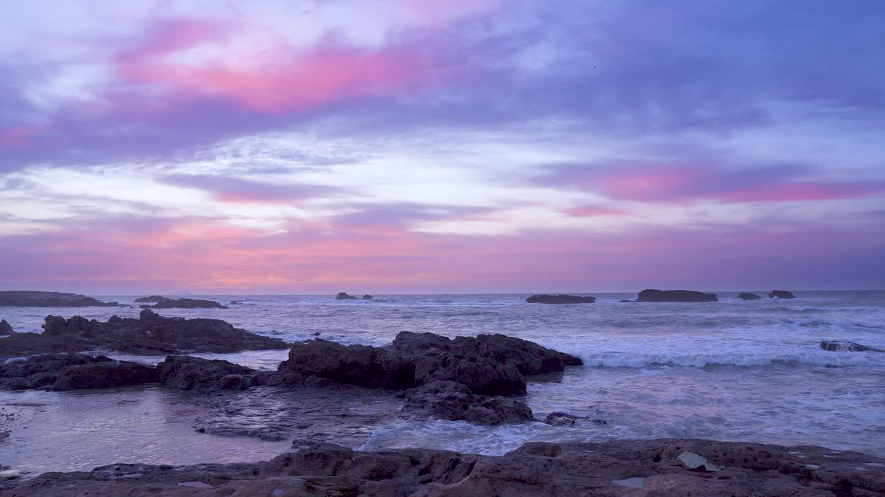 Purple colored clouds over ocean waves at sunset. Rocks and sand in the waters of the Atlantic Ocean. Seagull flying over water