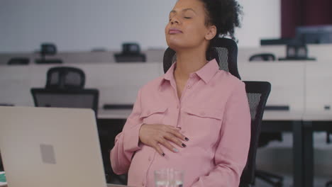 happy pregnant woman caressing her belly while sitting at desk in the office