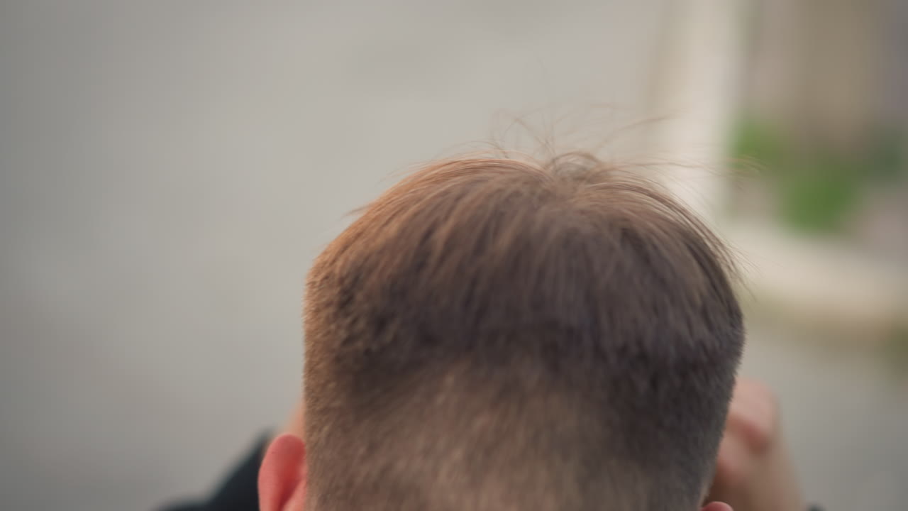 젊은 남자가 위에서 내려다보며 머리 왕관과 두피를 확인하고, 손가락으로 짧은 갈색 머리를 정돈하며, 머리 뿌리와 질감을 클로즈업하여 보여주고, 거울과 스타일링 준비를 암시하며, 조용한 촬영 전 일상, 자연광