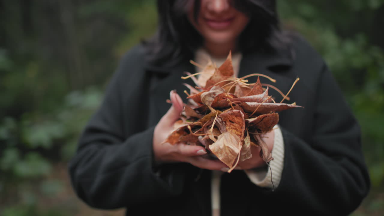 Hand view of beautiful young girl holding heap of dry autumn leaves, lifting single leaf to nose to smell, soft focus forest background, black coat and striped sweater, calm seasonal mood