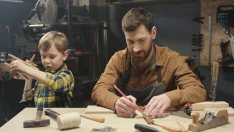 cheerful young caucasian carpenter working at desk in workshop and his little son playing next to him