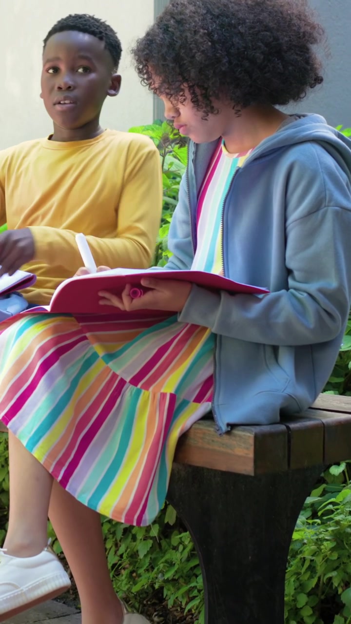 Vídeo vertical: Criança com blusa amarela e moletom azul folheando um fichário, escrevendo em uma carteira escolar enquanto estuda.