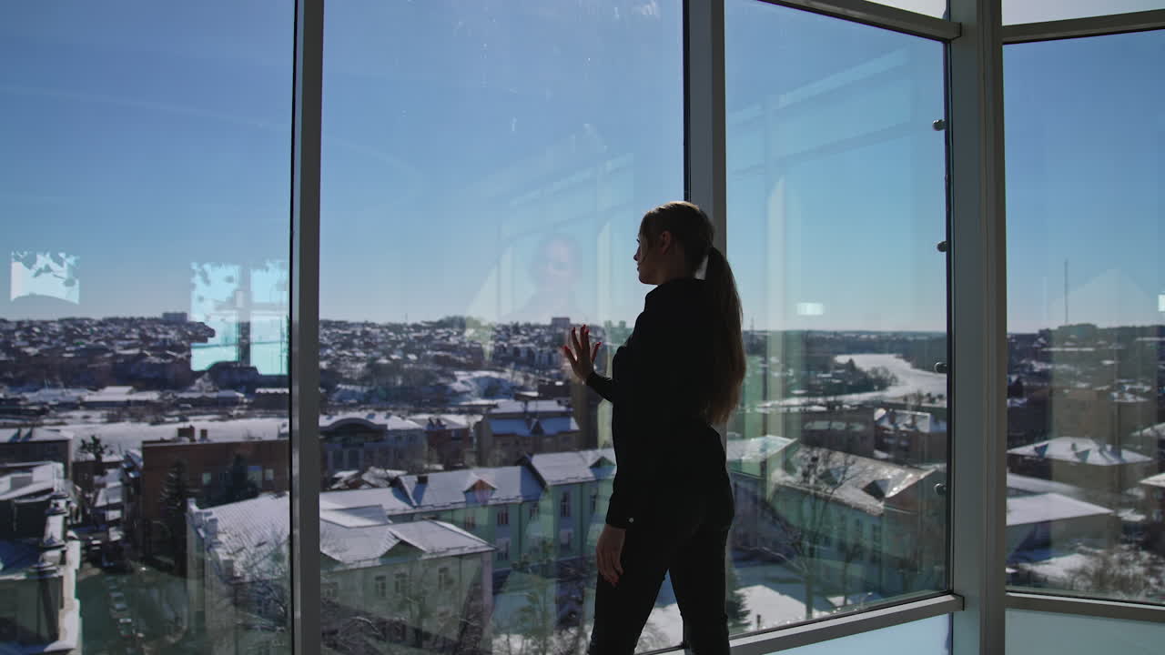 Young good-looking female wearing dark clothes standing in front of the panoramic windows. Sunrays coming into the big office room. Girl looking out of the window and enjoying the cityscape.