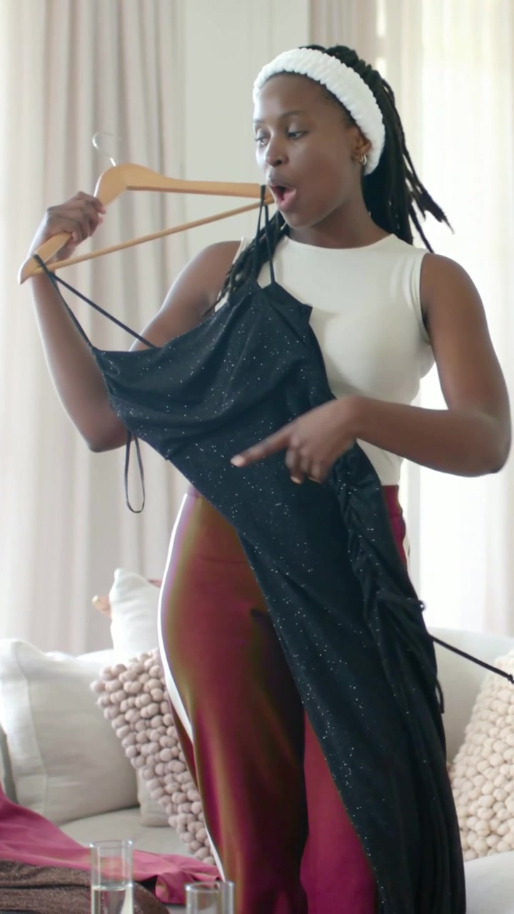 Vertical video: Lifting hanger, woman wearing headband holding black dress at home, checking fit