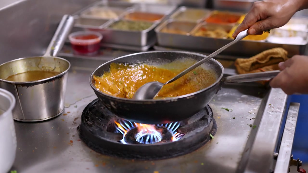 Kok hælder sauce og rører gul karry på en pande over blå gasflammer