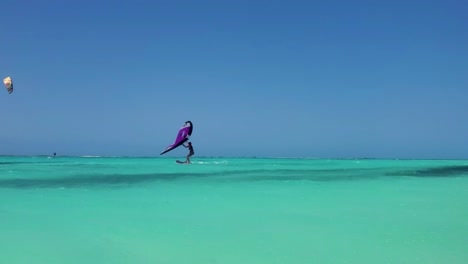 Woman-sailing-wing-foil-kite-on-crystal-caribbean-sea-and-man-kitesurfing-jump-behind