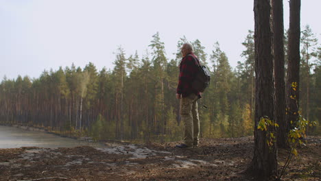 lonely fisherman is viewing forest lake from hill holding backpack and rod travelling in woodland at autumn day