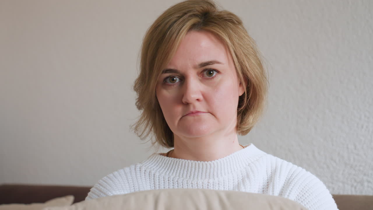 Woman with short hair and white sweater seated in therapy room holding beige pillow, gazing thoughtfully to side, calm expression on face, quiet atmosphere of emotional session