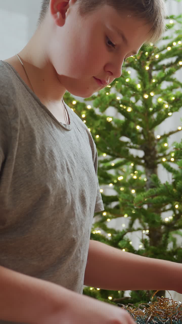 트리 옆에서 장식을 정리하는 소년, 장신구와 얇은 금속 조각을 놓고 전등을 풀면서 집중하는 표정, 캐주얼한 회색 셔츠, 작은 장식물을 살펴보는 손, 소나무 가지, 아늑한 집 분위기, 시즌
