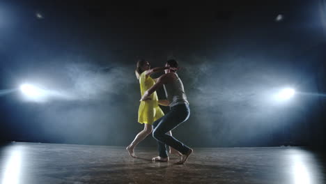 una joven con un vestido amarillo y un hombre están bailando rápido corriendo el uno hacia el otro y haciendo trucos acrobáticos en cámara lenta en el escenario en el humo y los reflectores