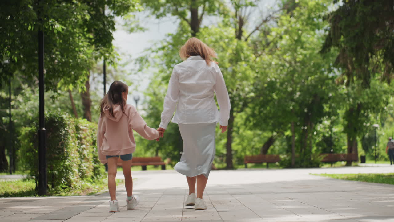 Moeder en dochter lopen door het park, ruggelings, hand in hand op een schaduwrijk pad, een langzame wandeling met een bedachtzame stemming. Jeansshorts, witte blouse, roze hoodie, bankje op de achtergrond, serene rust tussen generaties.