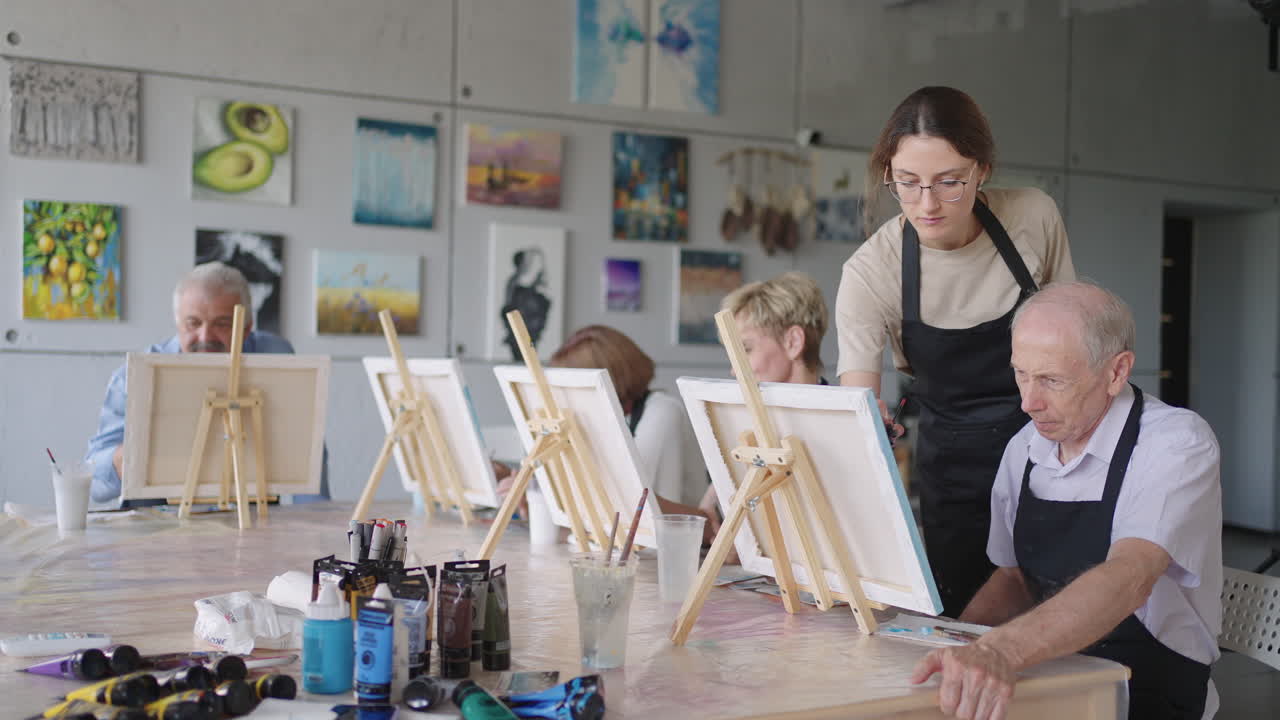 젊은 여성 교사가 은퇴자의 재활과 발전을 위한 과정에서 노인 학생들에게 아르티나를 그리는 데 도움을 주고 있습니다.