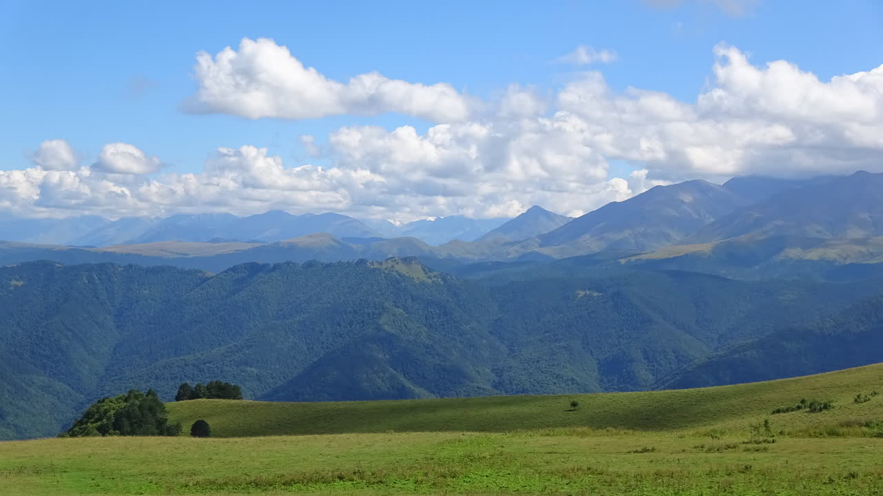 ロシアのコーカサス山脈の広角タイムラプス風景。手前には緑豊かな高山牧草地が広がり、重なり合う峰々の上には劇的な雲が流れています。