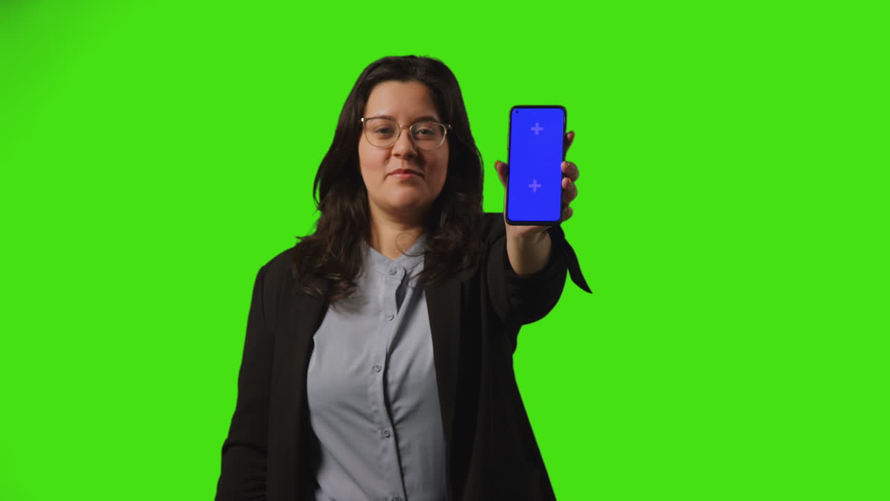 una joven mujer de negocios sonriente sosteniendo un teléfono móvil de pantalla azul hacia la cámara de pie contra el fondo de la pantalla verde