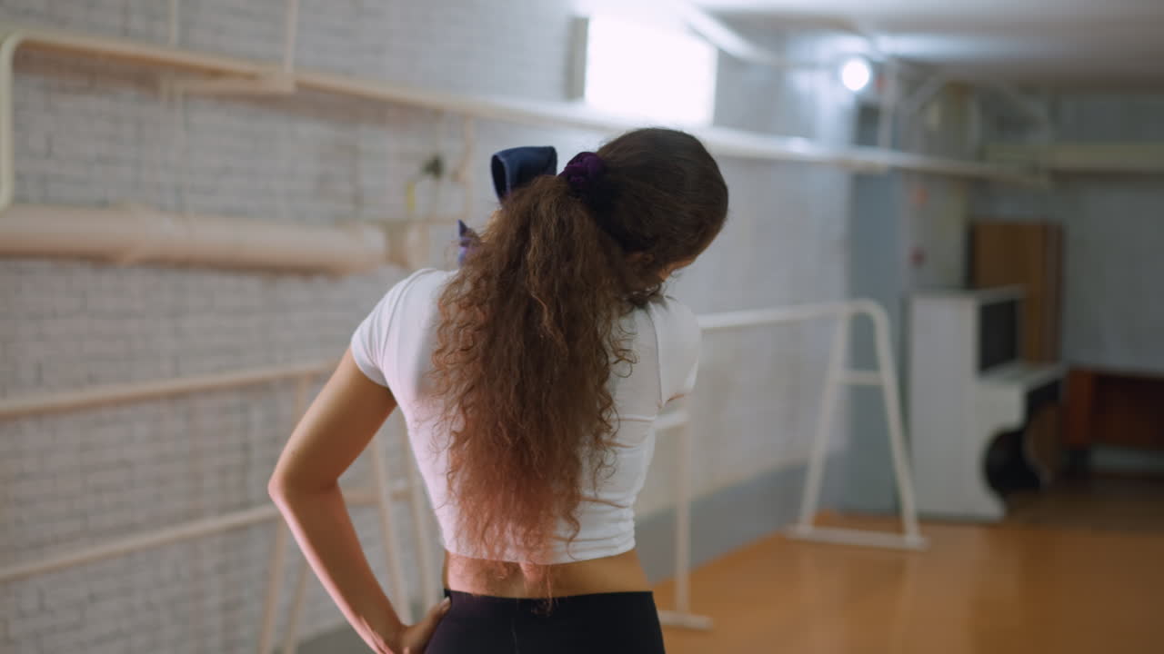 Back view of young woman in white top and black leggings placing hand on waist while wiping sweat from neck with blue towel after workout in fitness studio showing tiredness and relief from exercise