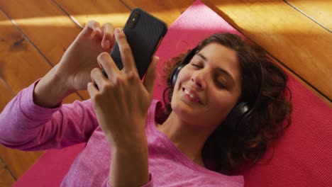 Feliz-Mujer-De-Raza-Mixta-Con-Auriculares,-Tumbada-En-Una-Estera-De-Yoga-Usando-Un-Teléfono-Inteligente-Y-Sonriendo-En-Una-Habitación-Soleada