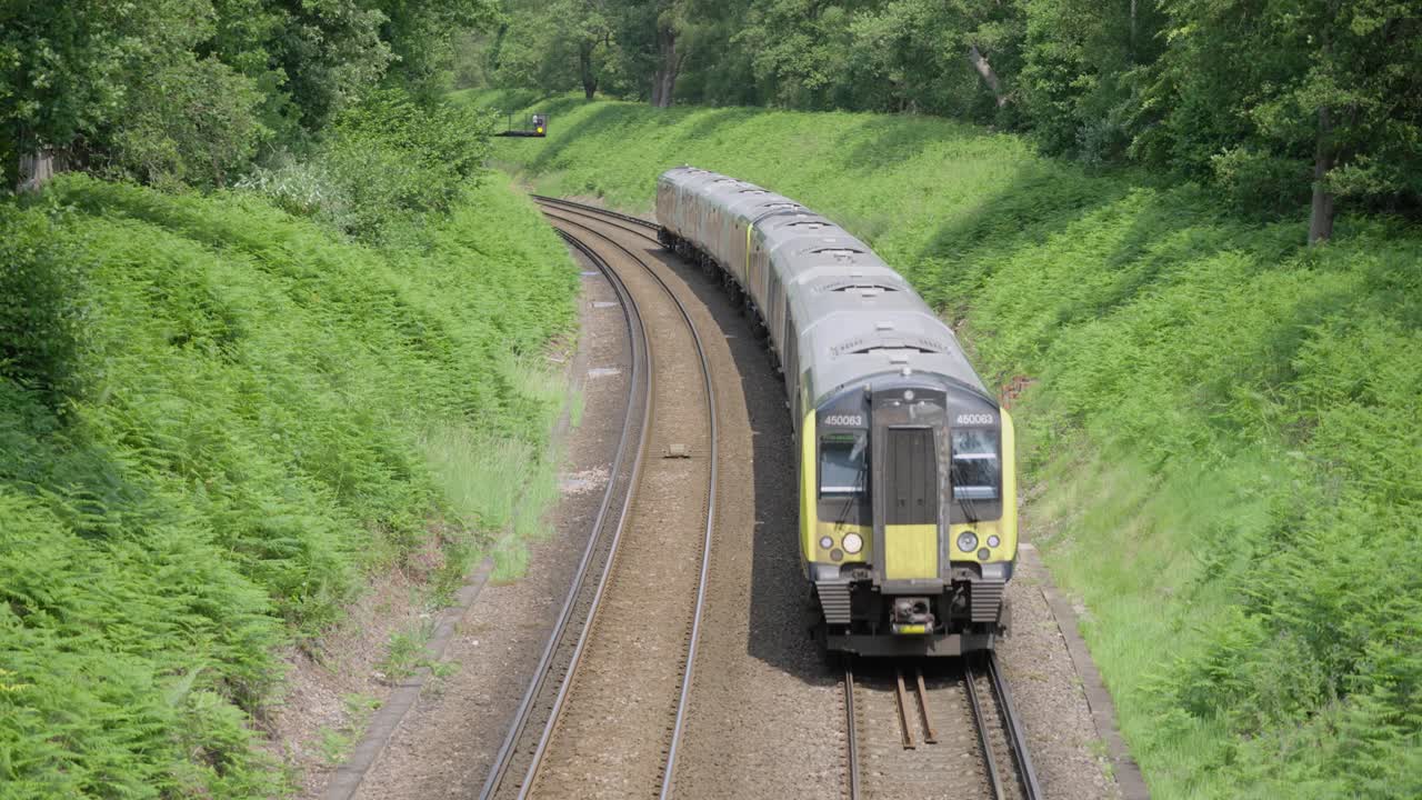 A modern British commuter train departs through green countryside on a curved double-track railway, surrounded by summer foliage and forest under an overcast sky.