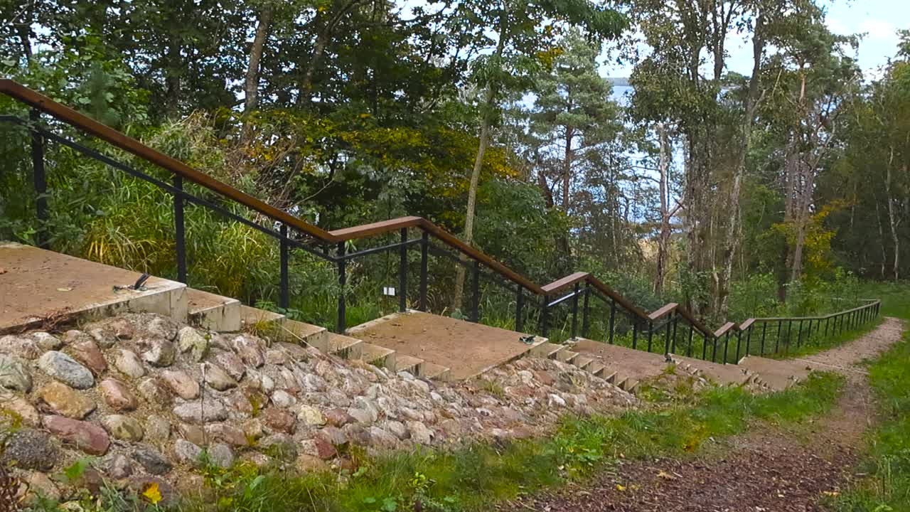 Naturskøn stentrappe, der fører ned gennem frodige grønne træer mod sandstrand ved Østersøen og kystklipper i Tabasalu, Estland.