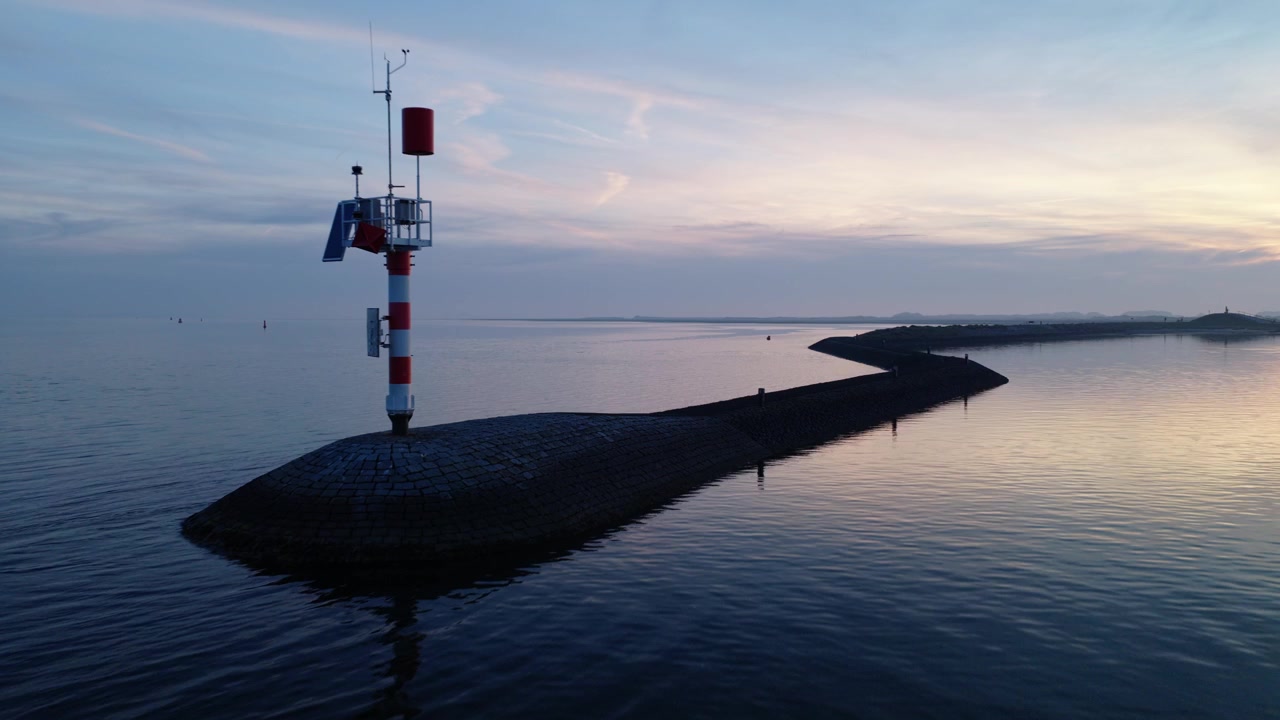 Drönare flyger bort från ett sjömärke i Vadehavet nära Terschelling, Friesland, Nederländerna, och avslöjar stenpirar, lugnt tidvatten och fridfull kustutsikt i blå timmen.