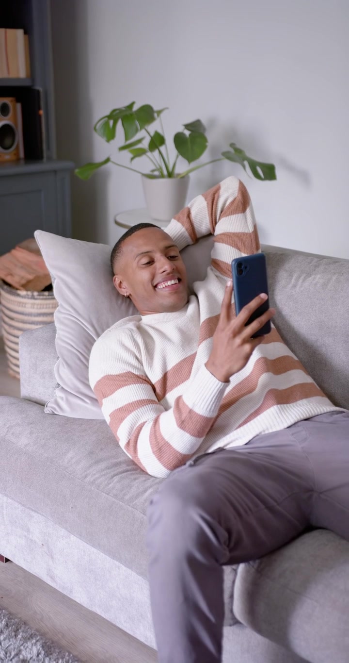 Video verticale: Uomo afroamericano sui venticinque anni sdraiato sul divano con un maglione, che scorre il telefono, sorridendo
