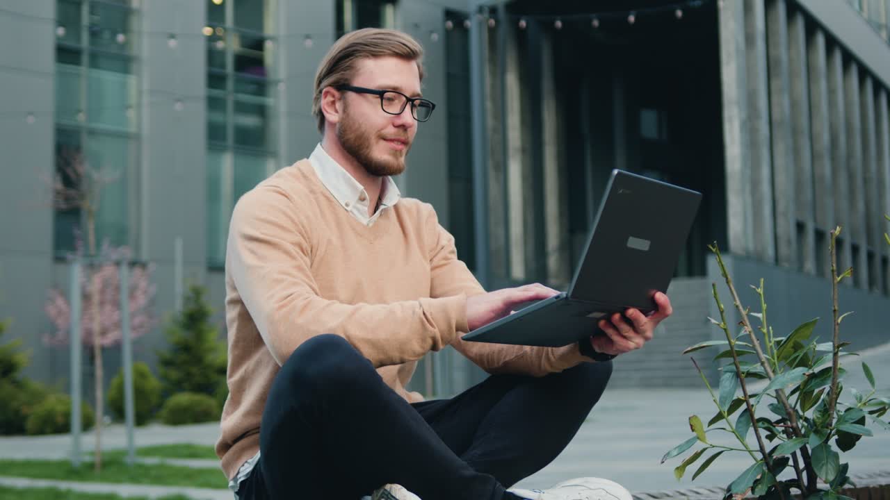 Handsome young caucasian male working or studying using laptop while sitting crossing legs on bench of city financial district. Outdoors