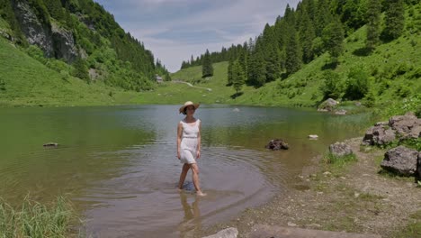 a woman in a white dress wades in talalpsee lake, surrounded by lush greenery and mountains