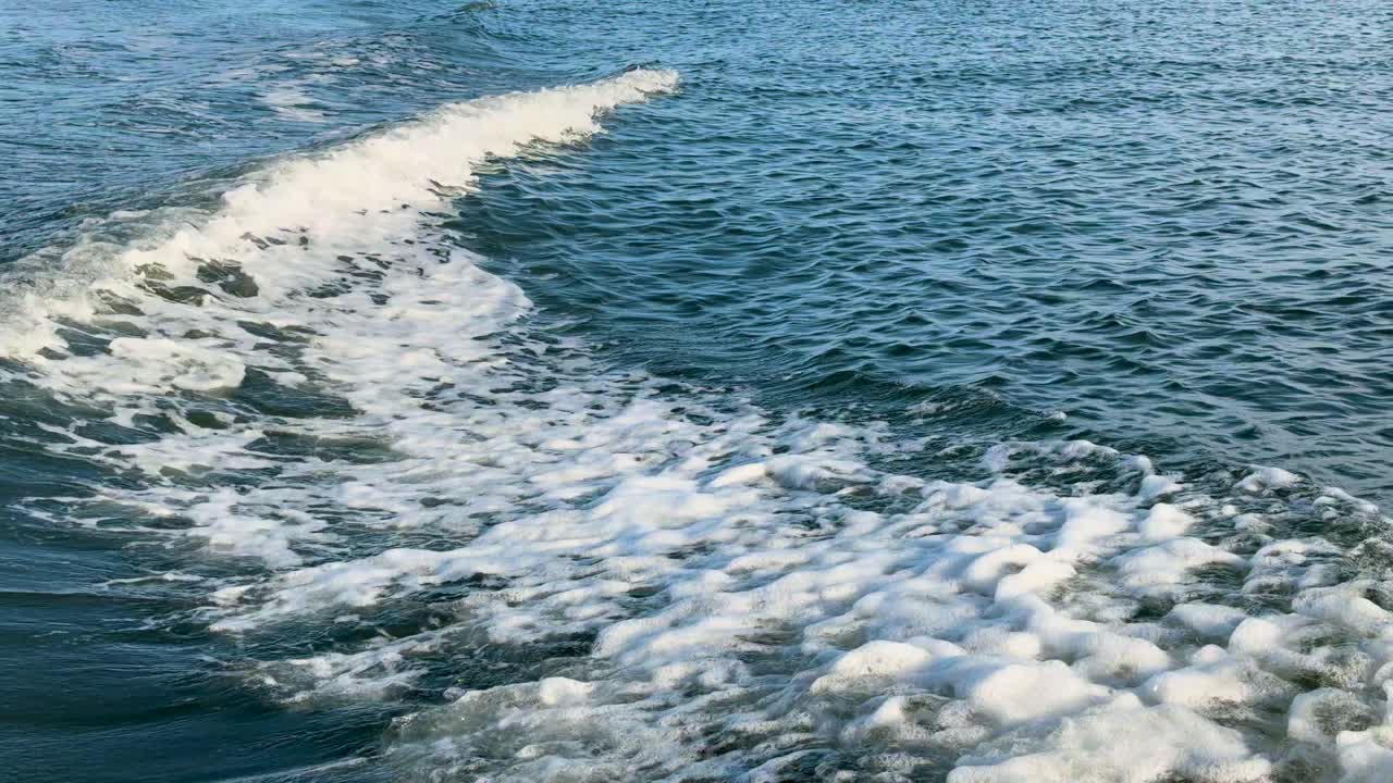 Foamy boat wake moves through blue ocean water, daylight, steady rear-facing camera, tranquil mood