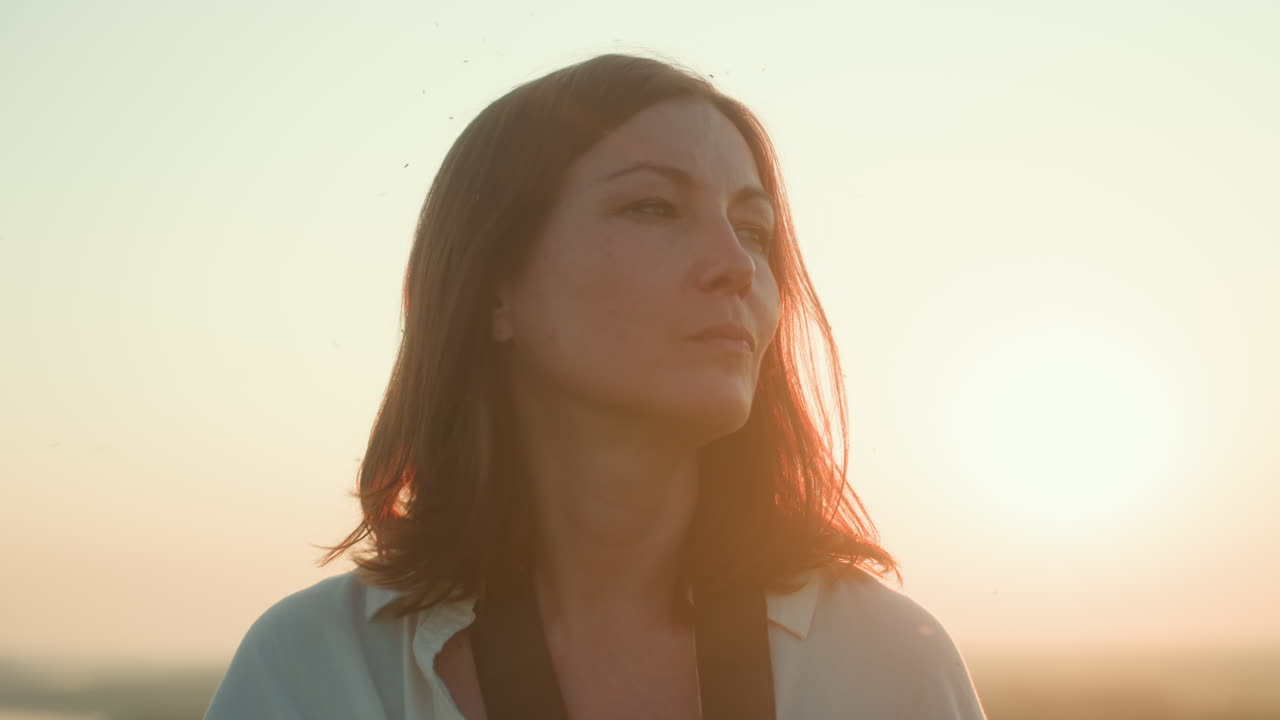 Close up of woman taking photograph during golden hour with warm sunlight casting bright glow around lens, soft smile as she captures moment in serene outdoor landscape filled with natural light