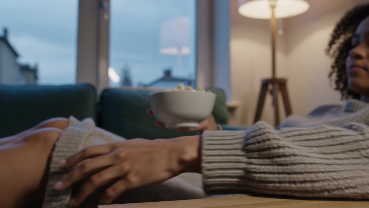 Accomodandosi, una donna afroamericana allunga la mano, prendendo popcorn da sgranocchiare a casa, indossando un maglione di lana
