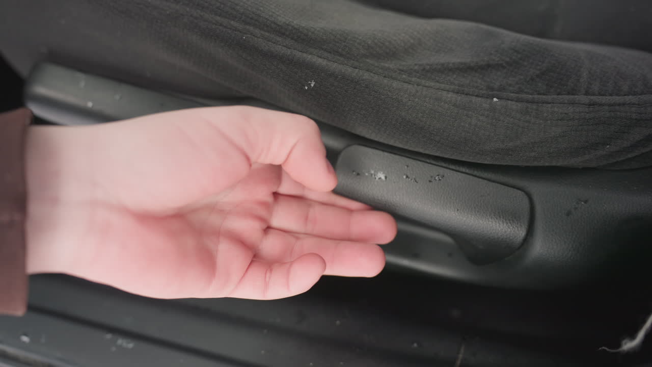 close up person hand holding car seat lever while adjusting seat backward on snowy day with visible snowflakes on seat fabric and matte black handle inside vehicle interior during winter