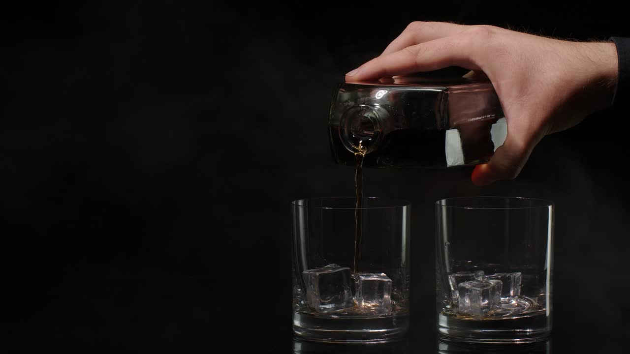 Barman versa whisky dorato, cognac e brandy dalla bottiglia nei bicchieri con cubetti di ghiaccio su sfondo scuro.