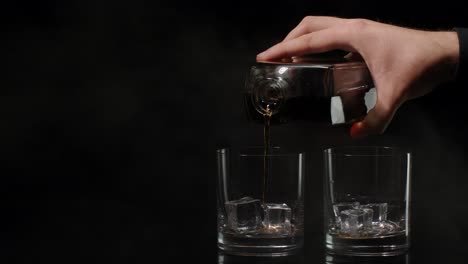 barman pour golden whiskey cognac brandy from bottle into glasses with ice cubes on dark background